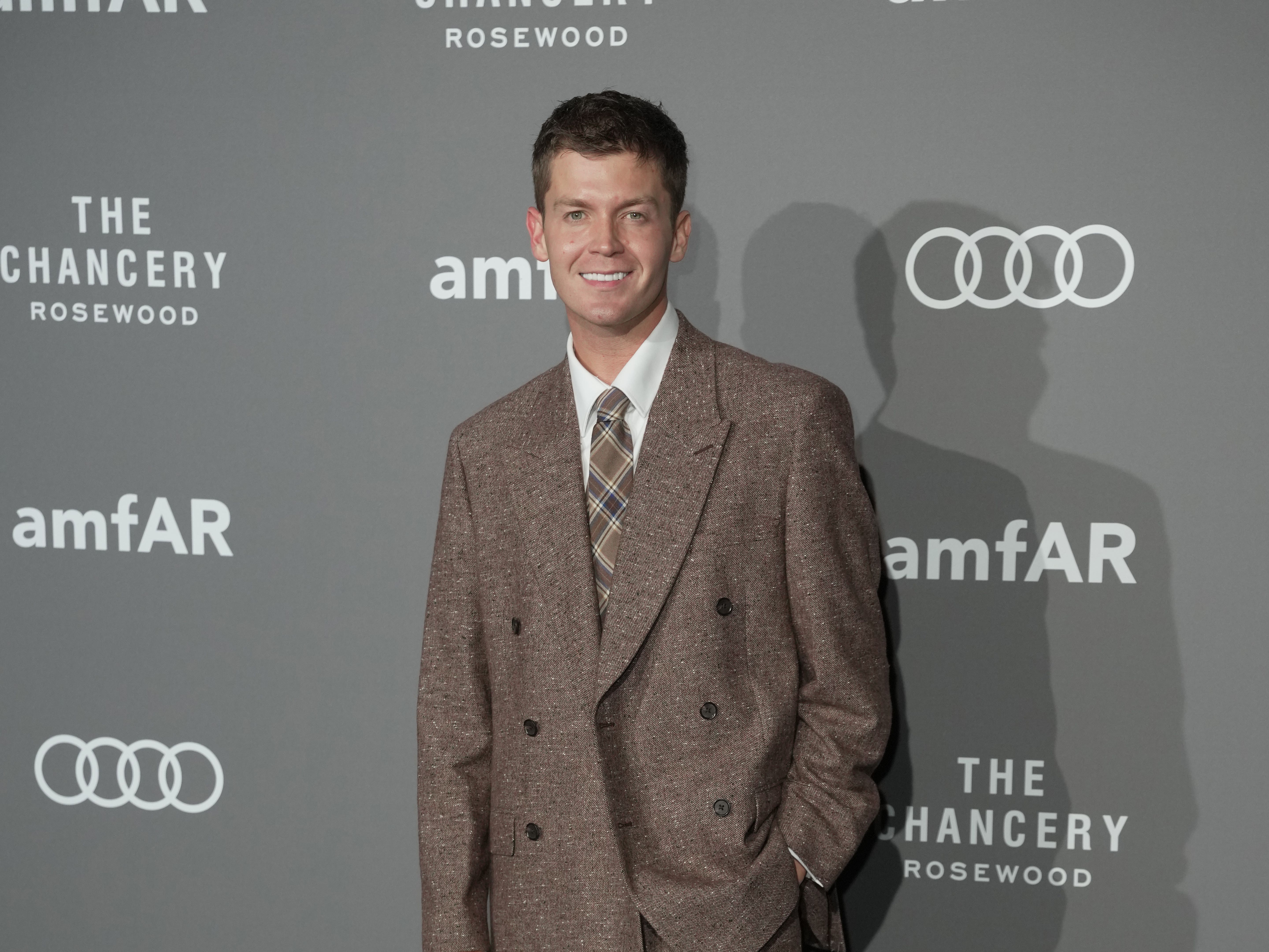 Jamie Miller smiling while posing for photos at a red carpet event, wearing a brown suit